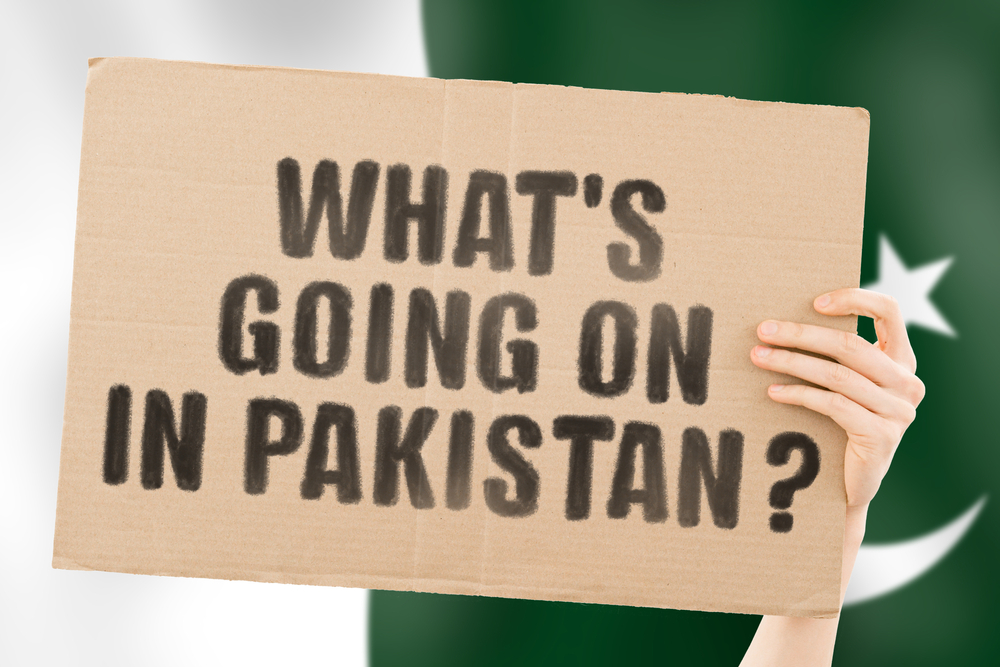 The question " What's going on in Pakistan? " on a banner in men's hand with blurred Pakistani flag on the background. Protests on the street. Riot. Violence. Economic crisis. Collapse. Politics
