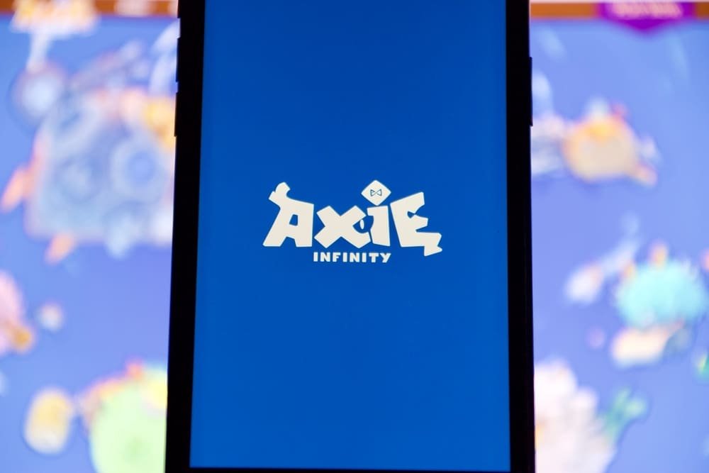 Aug 27 2021-Lopburi, Thailand: Axie blue color logo on smart mobile phone screen, blur laptop computer with characters on screen as a background. Axie Infinity is NFT game using play to earn concept.