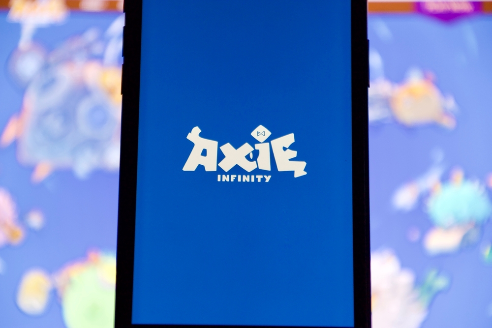 Aug 27 2021-Lopburi, Thailand: Axie blue color logo on smart mobile phone screen, blur laptop computer with characters on screen as a background. Axie Infinity is NFT game using play to earn concept.