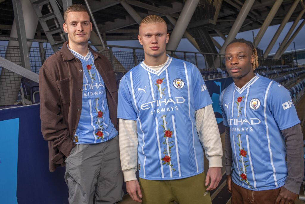 Manchester City stars Sergio Gómez, Erling Haaland, and Jérémy Doku model the "The Roses and the Bees" jerseys with designs by artist Christian Jeffery.






